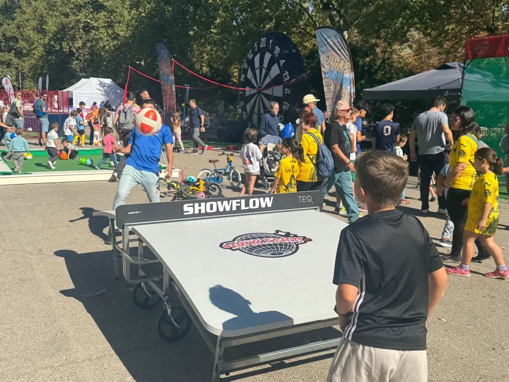 Grupo de niños y adultos reunidos alrededor de una mesa triangular de juego llamada SNOW-LOW, en un evento al aire libre con árboles y carpas. La mesa tiene logotipos visibles y parece diseñada para deslizar o lanzar objetos.