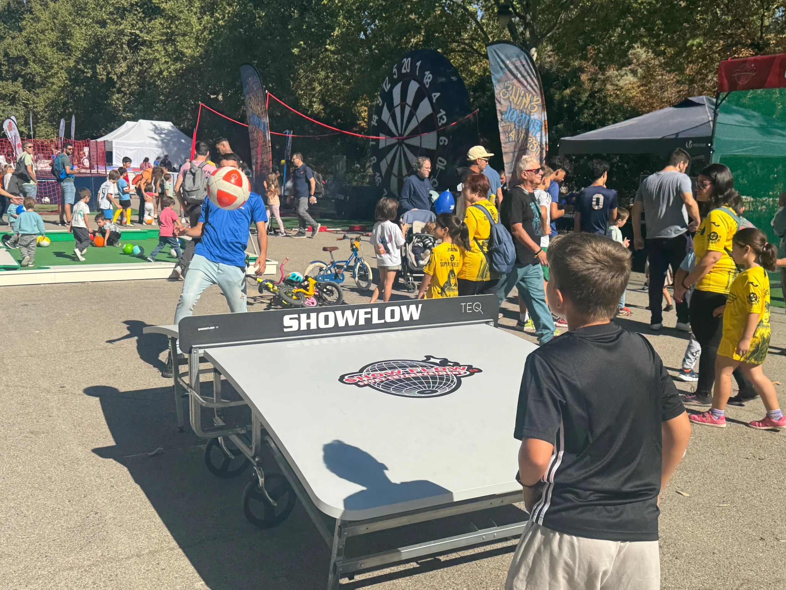 Grupo de niños y adultos reunidos alrededor de una mesa triangular de juego llamada SNOW-LOW, en un evento al aire libre con árboles y carpas. La mesa tiene logotipos visibles y parece diseñada para deslizar o lanzar objetos.