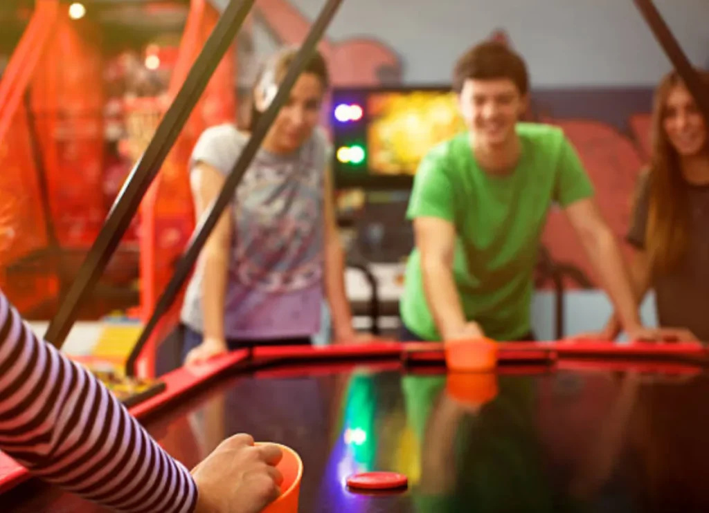 Dos personas jugando futbolín en área recreativa bien iluminada. No hay mesa de air hockey en esta imagen.
