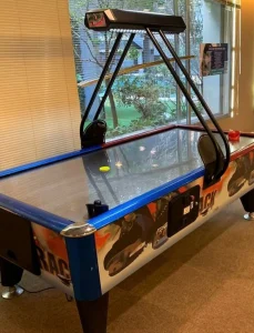 Mesa de air hockey con estructura de madera y superficie azul, colocada en habitación con piso de madera.