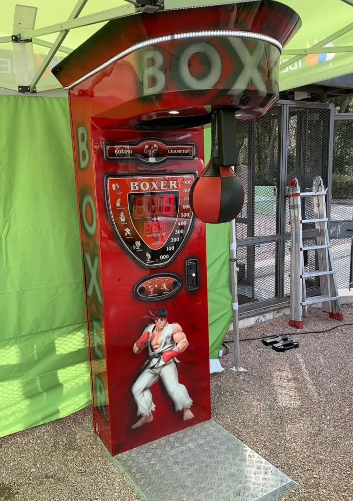Máquina recreativa de boxeo roja con un dibujo de un boxeador, instalada al aire libre.