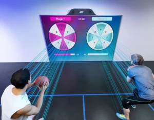 Dos hombres compitiendo en un simulador de pared interactivo MultiBall con un juego de ruleta o diana proyectado. Cada jugador lanza un balón de baloncesto para impactar en su ruleta de puntuación (Player 1 en magenta y Player 2 en cian).