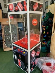 Máquina de garra con temática Coca-Cola ubicada en un espacio interior recreativo.
