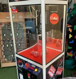 Máquina de garra con temática Coca-Cola ubicada en un espacio interior recreativo.