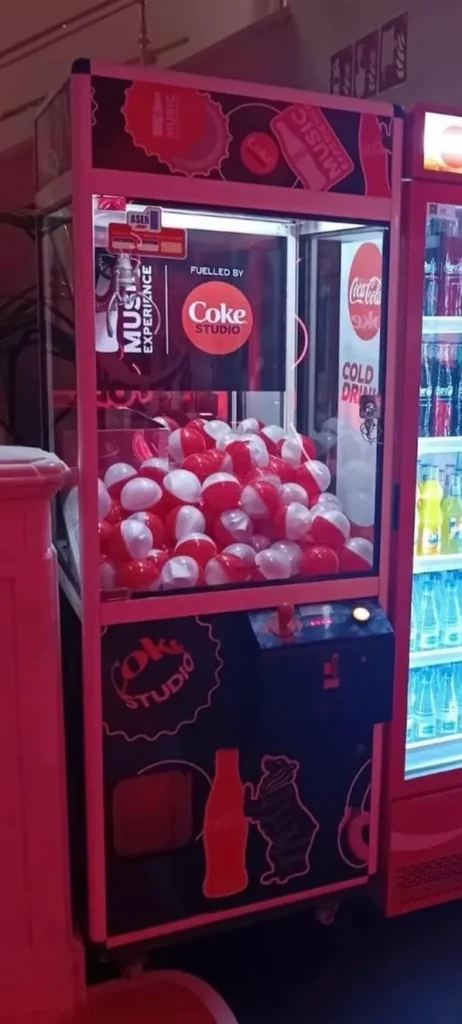 Máquina de garra con temática de Coke Studio, ubicada junto a un refrigerador de bebidas embotelladas.