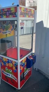 Máquina de garra con temática de Coke Studio, ubicada junto a un refrigerador de bebidas embotelladas. Contiene bolas plásticas rojas y blancas como premios.