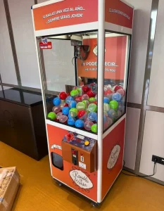 Máquina de garra ubicada en interior junto a pared con paneles de madera y toma eléctrica.