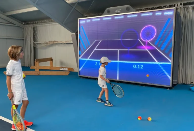 Simulador de tenis MultiBall para niños. Dos jugadores en cancha azul con pantalla de juego interactiva.