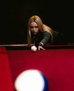 Mujer joven con cabello rubio preparándose para golpear una bola en una mesa de billar de paño rojo.