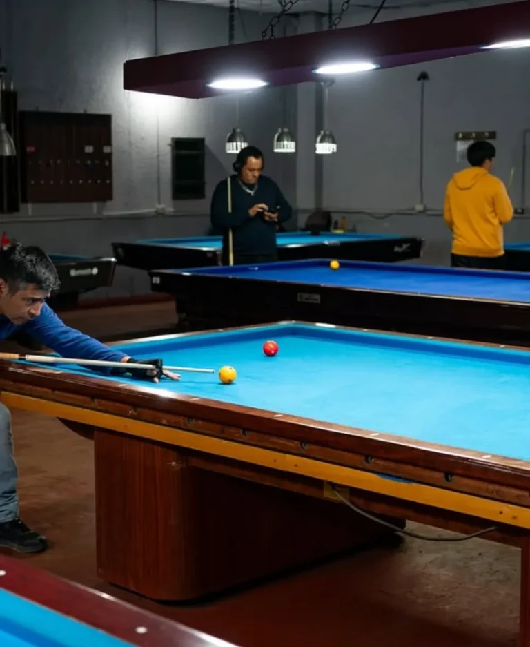 Dos personas jugando billar en una mesa clásica de paño marrón en un salón interior oscuro.