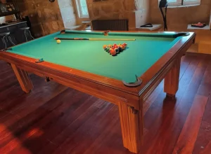 Mesa de billar de madera rojiza y paño verde en un suelo de madera.