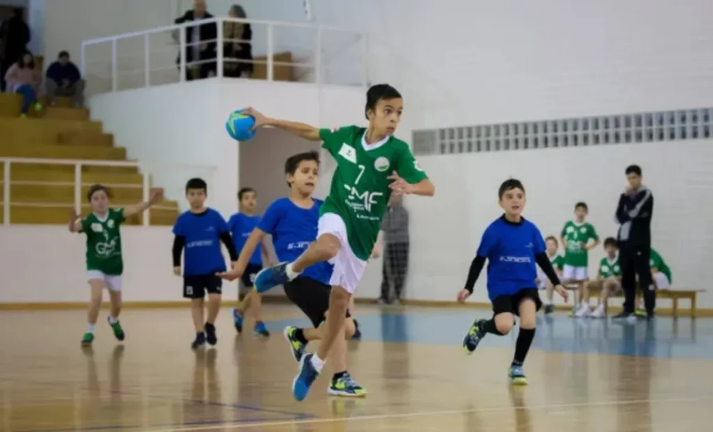 Jóvenes jugadores de balonmano en acción durante un partido en pabellón, con un niño en primer plano botando el balón mientras sus compañeros y monitores observan al fondo.