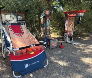Dos estaciones de máquinas de baloncesto arcade (basket shoot) situadas al aire libre en una zona de tierra rodeada de árboles para una jornada de convivencia.