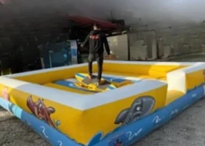 Atracción de tabla de surf mecánica con base inflable amarilla y azul, capturada en un ángulo amplio durante un montaje en exteriores.