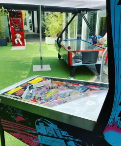 Primer plano de una mesa de pinball clásica con gráficos coloridos y luces encendidas, instalada bajo una carpa blanca en un evento recreativo.