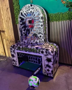 Máquina recreativa de "golpe de fútbol" con diseño de portería y panel luminoso de puntuación, ubicada en un espacio interior decorado con estilo moderno.
