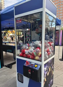 Máquina de garra atrapa-peluches (claw machine) profesional con estructura blanca y azul, llena de premios y lista para el uso del público en una feria.