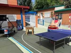 Mesa de ping-pong (tenis de mesa) profesional instalada en una pista deportiva exterior junto a otras actividades recreativas bajo el sol.