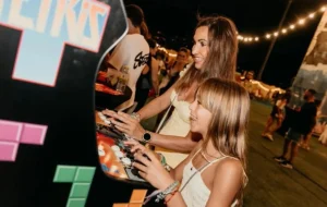Dos mujeres jóvenes, con pelo rubio, sonriendo y jugando juntas en la consola de control de una máquina recreativa en un evento nocturno al aire libre.