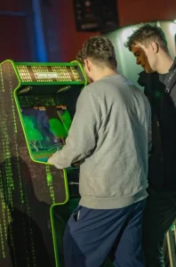 Dos hombres jóvenes, vistos desde atrás, jugando en una máquina recreativa clásica iluminada con luces verdes de neón en un entorno oscuro.
