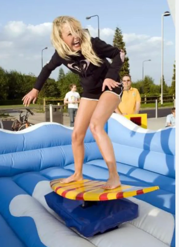 Mujer joven rubia parada sobre un simulador de surf mecánico de color amarillo y rojo con diseño de tabla de madera, que se encuentra dentro de una colchoneta inflable azul. Viste ropa negra de surf.
