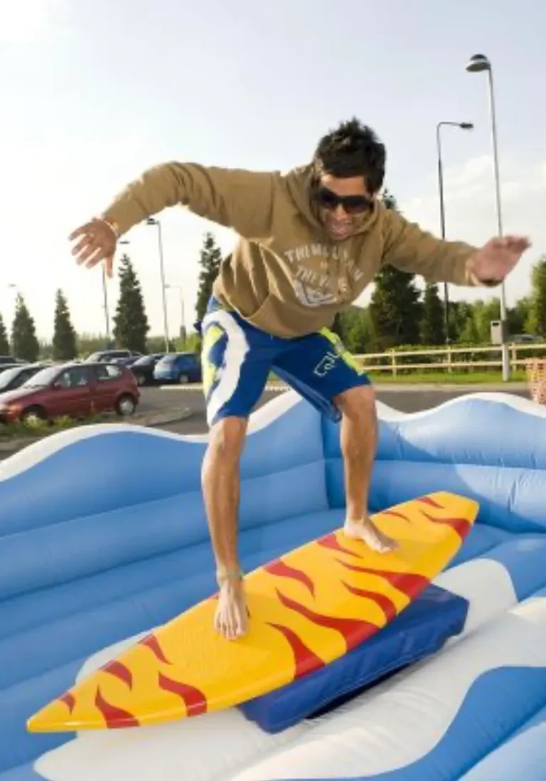 Hombre con gafas de sol, sudadera marrón y shorts de surf azules, sonriendo y manteniendo el equilibrio descalzo sobre un simulador de surf mecánico de color amarillo y rojo con diseño de llamas. El inflable de protección es de color azul y blanco, imitando olas.