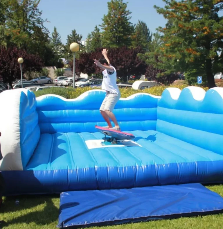 Hombre joven con camiseta blanca saltando o cayendo de un simulador de surf o skate mecánico, cayendo sobre la colchoneta inflable azul. El evento se realiza al aire libre en un entorno verde.