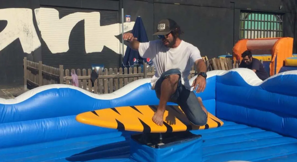 Hombre joven con gorra y gafas de sol, vistiendo shorts de baño, intentando mantener el equilibrio en un simulador de surf mecánico con una tabla amarilla. El juego está rodeado por un inflable azul.