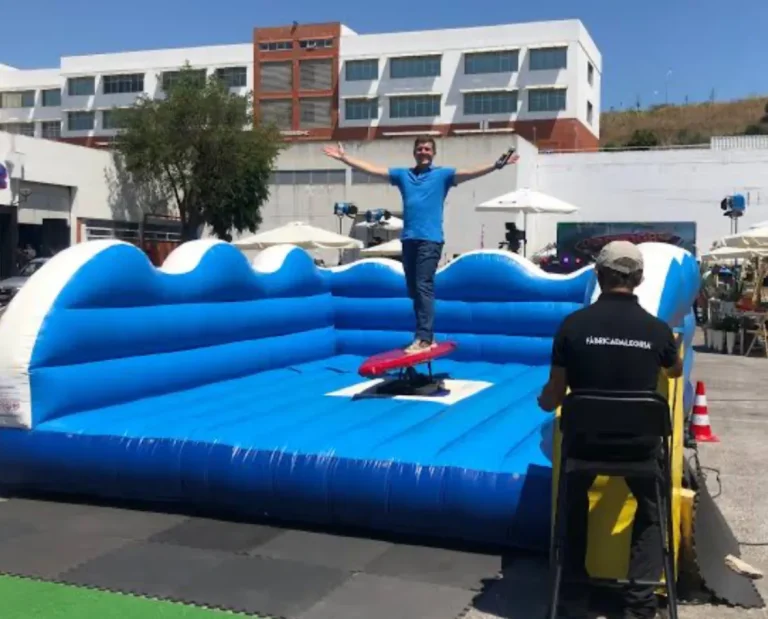 Hombre con camiseta azul y jeans manteniendo el equilibrio con los brazos abiertos sobre un simulador de surf mecánico de color rojo. El juego está dentro de un inflable grande de color azul con forma de olas, al aire libre en una plaza o estacionamiento. Un operario vestido de negro está en el panel de control.
