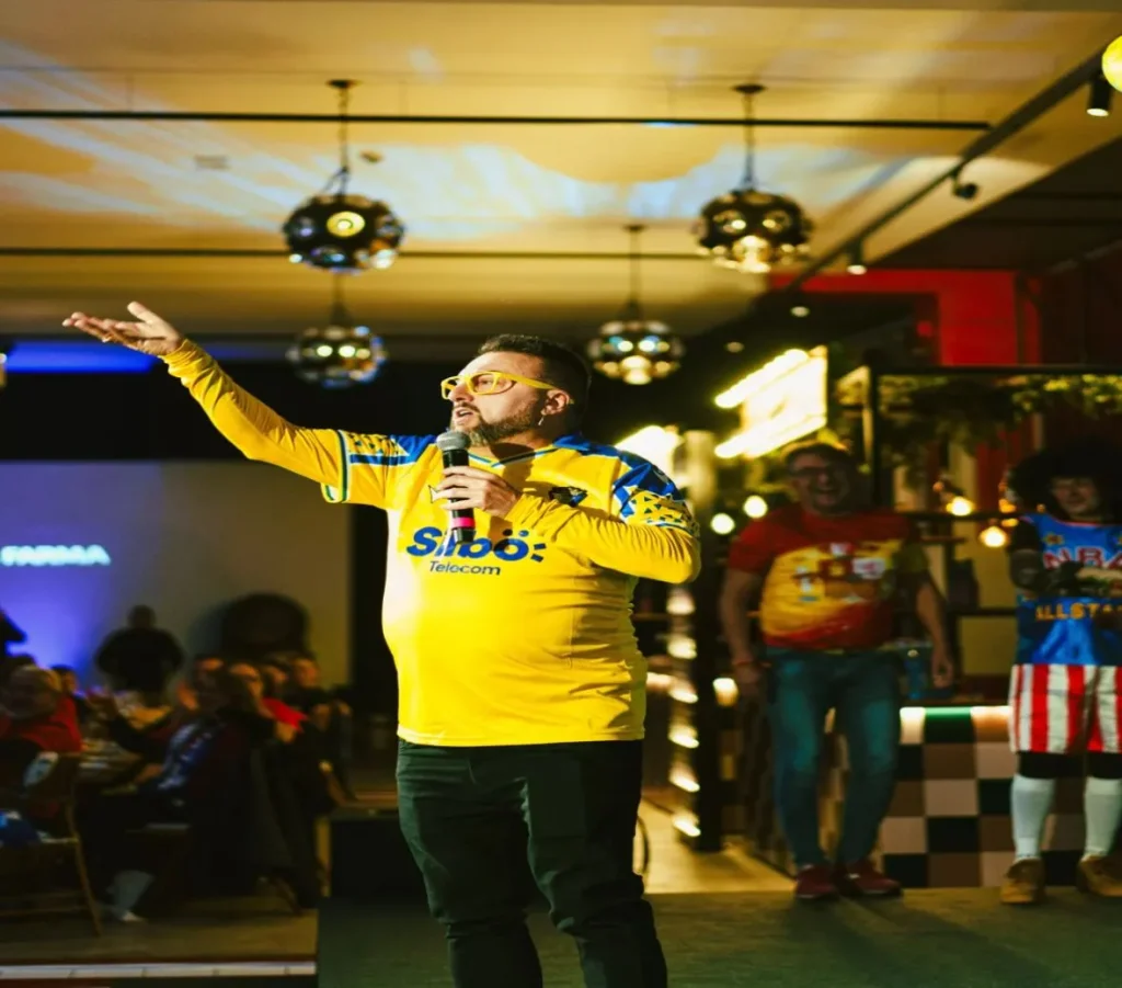 Speaker de eventos corporativos o deportivos hablando en un escenario indoor con luces de fiesta.