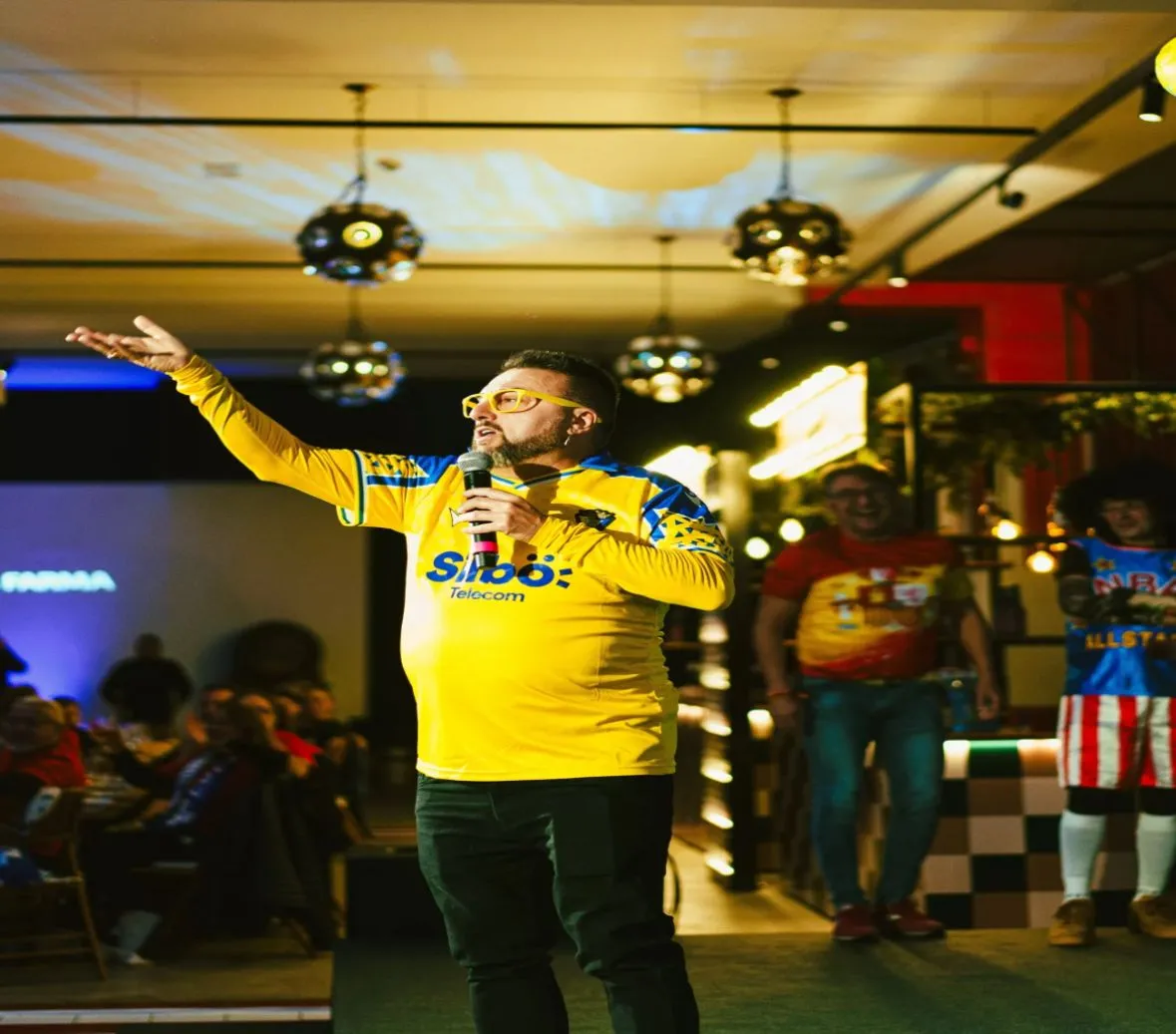 Speaker de eventos corporativos o deportivos hablando en un escenario indoor con luces de fiesta.