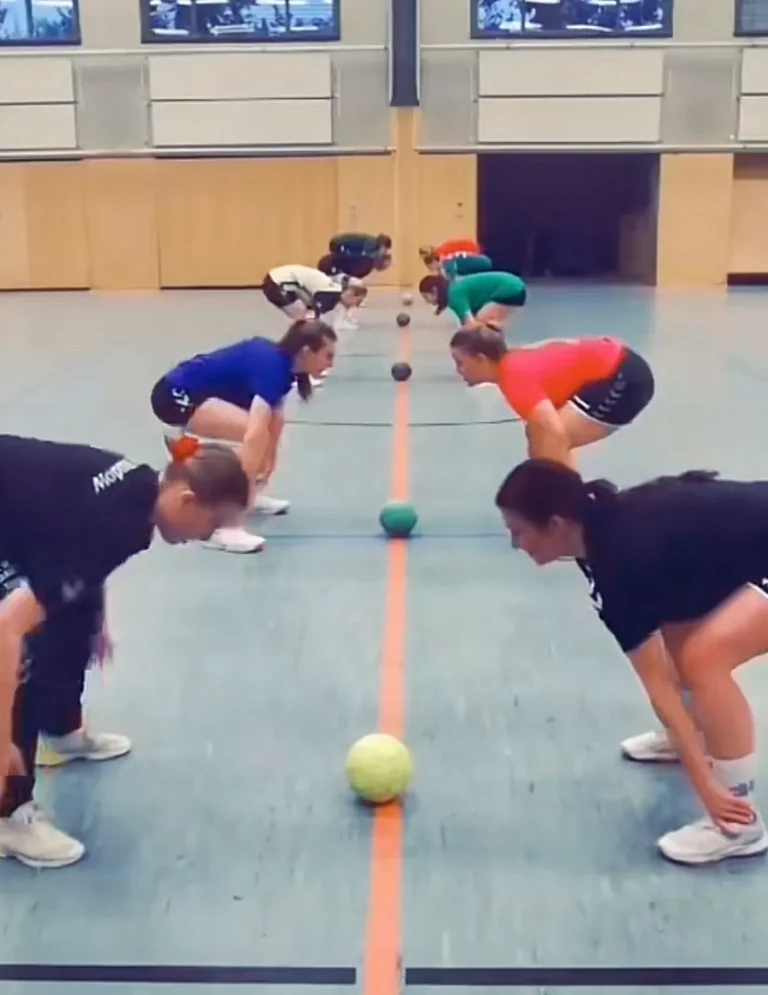 Grupo de niños agachados y alineados en una cancha, con una línea naranja en el suelo, participando en un ejercicio de entrenamiento de balonmano o educación física con pelotas pequeñas.