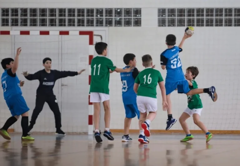 Varios niños con uniformes de balonmano (algunos verdes, otros azules) parados juntos en una formación defensiva u ofensiva en la cancha de un pabellón deportivo.