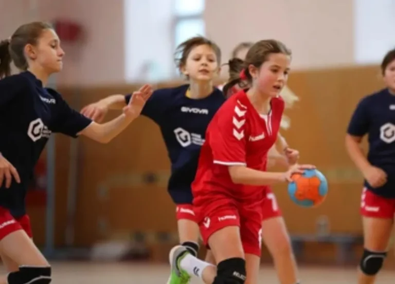 Tres niñas o adolescentes jugando balonmano. Una jugadora con una camiseta negra sostiene una pelota naranja y se prepara para pasarla o lanzar, mientras otras dos jugadoras la miran.