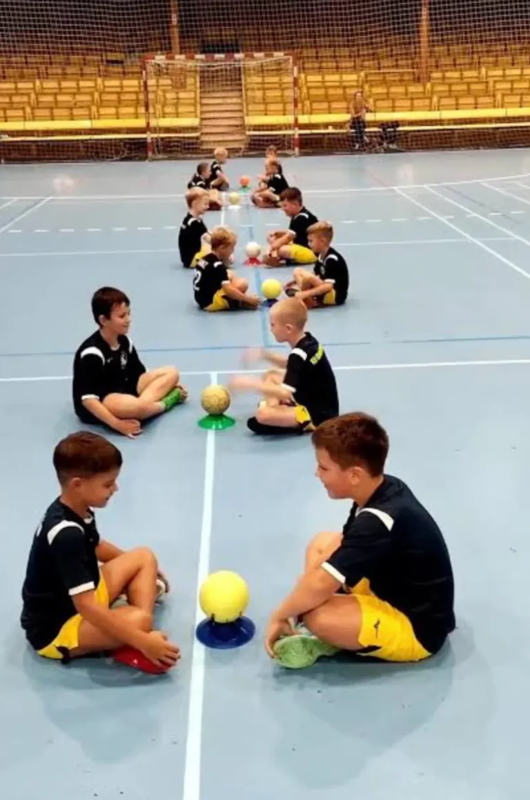 Grupo de niños sentados en el suelo de un pabellón deportivo, participando en un juego o actividad de balonmano con pelotas pequeñas y conos de colores.