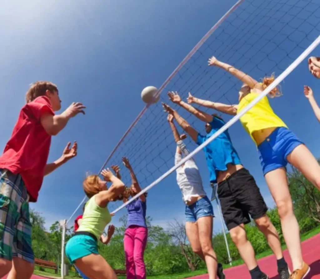 Un equipo de jóvenes celebrando un punto en una pista al aire libre, con la red de voleibol de fondo y un cielo despejado que resalta el carácter festivo de la actividad.