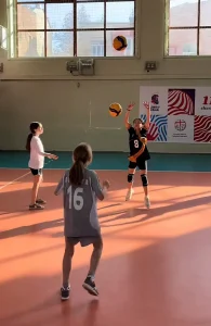 Dos niñas en una pista polideportiva practicando el saque de voleibol bajo la supervisión de monitores, con iluminación natural que destaca la amplitud del espacio.