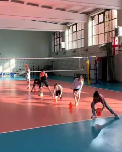 Vista amplia de una sesión de entrenamiento de voleibol en pabellón, con varios grupos de niños realizando ejercicios dinámicos de desplazamiento y control de balón sobre una pista azul y roja.