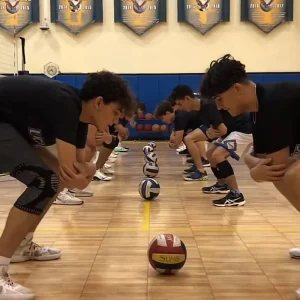 Un grupo de niños alineados en posición de defensa baja en la pista, concentrados en un ejercicio de agilidad y reflejos con balones de voleibol reglamentarios.