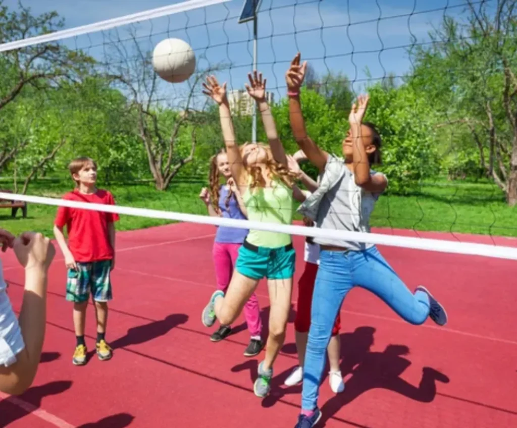 Jugadores de voleibol saltando junto a la red en una pista exterior roja durante un día soleado, capturando el momento del bloqueo en un ambiente recreativo.
