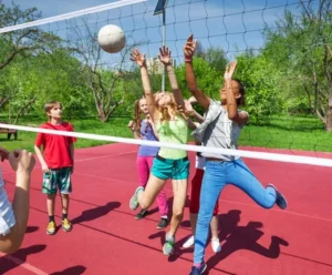 Jugadores de voleibol saltando junto a la red en una pista exterior roja durante un día soleado, capturando el momento del bloqueo en un ambiente recreativo.