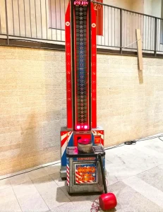 Una máquina de martillo de fuerza de feria instalada en un patio exterior; la torre vertical muestra luces rojas indicadoras y el mazo rojo descansa en el suelo junto a la base de golpeo.