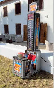 Vista lateral de la atracción de fuerza "High Striker" situada en un jardín exterior junto a un edificio blanco, destacando el diseño retro de la torre y el yunque de impacto.