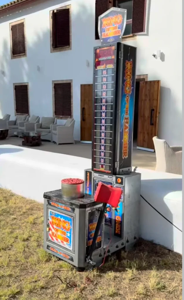 Vista lateral de la atracción de fuerza "High Striker" situada en un jardín exterior junto a un edificio blanco, destacando el diseño retro de la torre y el yunque de impacto.
