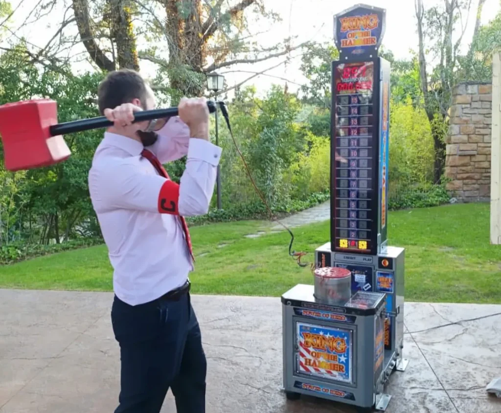 Un hombre con camisa blanca y corbata sujetando el mazo sobre sus hombros mientras se prepara para golpear la máquina de martillo en un evento al aire libre.