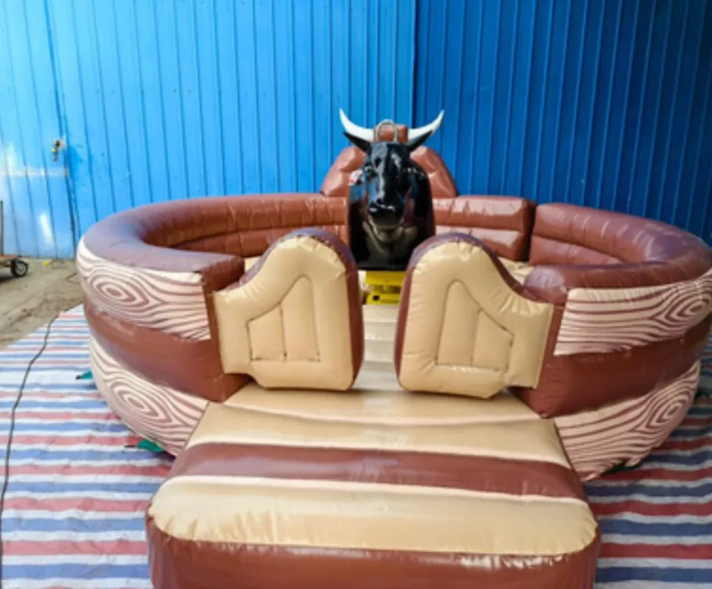 Toro mecánico de cuero marrón sobre una base inflable grande con dos sillones inflables de color claro, visto de frente contra un fondo azul.