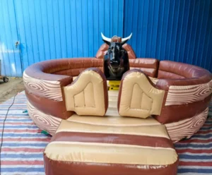 Toro mecánico de cuero marrón sobre una base inflable grande con dos sillones inflables de color claro, visto de frente contra un fondo azul.