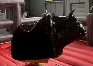 Primer plano de la parte trasera de un toro mecánico de color negro sobre una colchoneta inflable roja, probablemente capturado en interiores o en un stand.