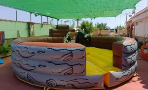 Vista aérea de un toro mecánico inflable rodeado por una colchoneta de protección amarilla y naranja con patrones de relámpagos negros, bajo una estructura de toldo.
