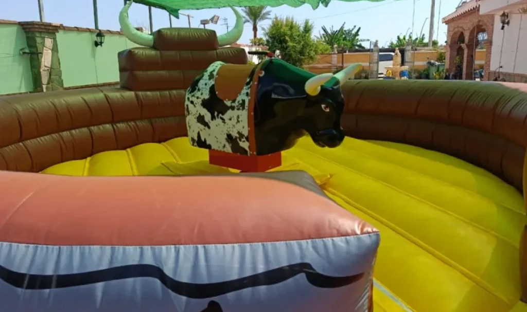 Toro mecánico de juguete con un jinete de dibujos animados sobre una base inflable de colores amarillo y marrón, en un entorno de patio con paredes de ladrillo.